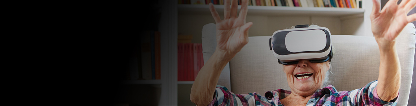 Eldre kvinne med VR-briller sitter i en lenestol og smiler med hendene i været.