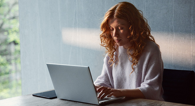 Ung kvinne sitter konsentrert og skriver på en laptop som ligger på skrivebordet foran henne. Inn fra siden kommer det solskinn som delvis skinner på kvinnens ansikt. Bak henne, en grå betongvegg.