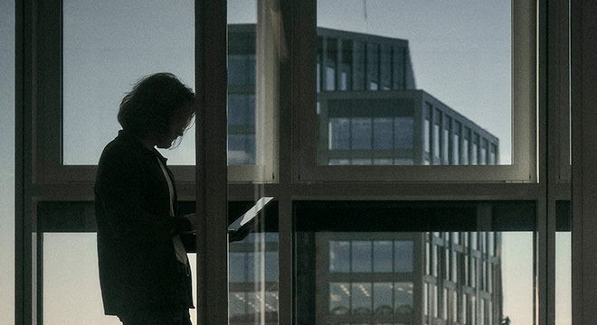 Mann står i profil med laptop i et dunkelt kontorlandskap, kveldshimmel og andre næringsbygg vises gjennom vinduene i bakgrunnen.