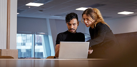 Mann og kvinne ved laptop i møterom. Mannen sitter ved pulten, kvinnen står ved siden av han. Begge ser på skjermen på laptopen.