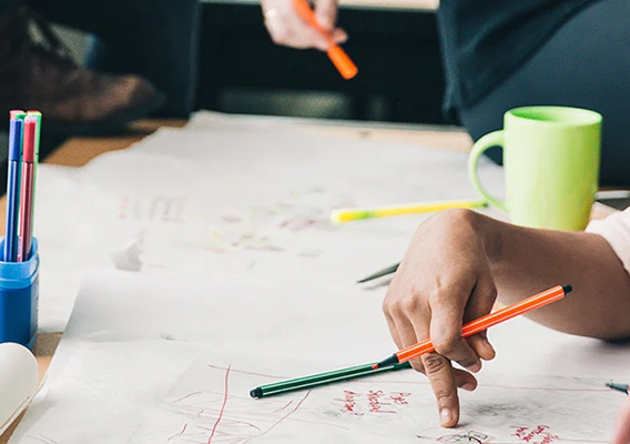 Skrivebord med papir, tusjer og lapper, hender som holder tusj og peker mot bordet. Kaffekopp.