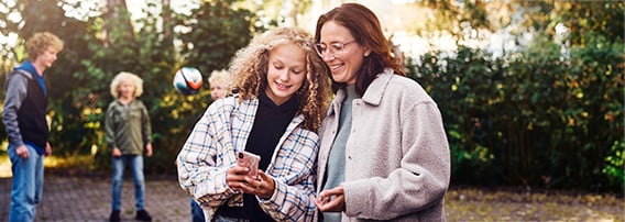 Høstkledt mor og barn i hagen, mor og datter står i forgrunnen og ser på en mobiltelefon mens de smiler.