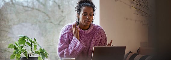 Kvinne iført ullgenser sitter ved kjøkkenbordet med kaffekopp og laptop.